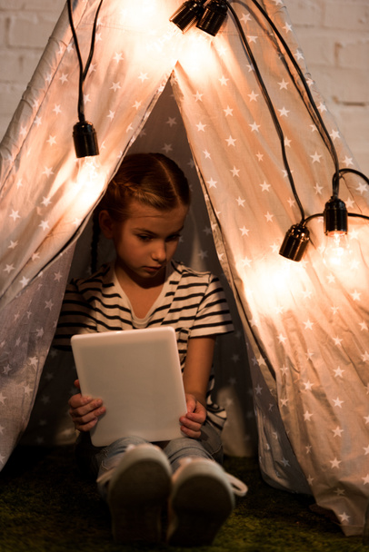 Niño serio sentado en wigwam y sosteniendo la tableta digital en la habitación oscura
 - Foto, imagen