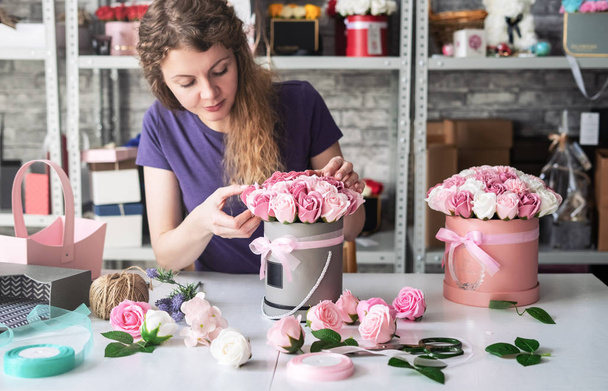 Flower shop: a florist girl collects a bouquet in a round box of pink roses. Blond curly hair, gray background. - Foto, afbeelding