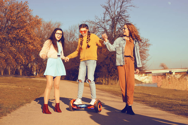 Trzy piękne dziewczyny w hoverboard. Relacji dziewczyny, spacerem, aktywne, nowe nowoczesne technologie. Dziewczyn uczyć dziewczyna jeździć w hoverboard. Słoneczny dzień, dziewczyny są zabawę, uśmiech, odkryty. - Zdjęcie, obraz