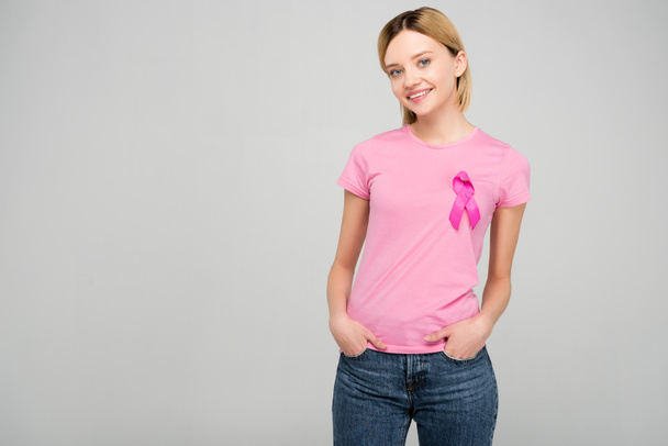 χαμόγελο ξανθό κορίτσι στο ροζ μπλουζάκι με κορδέλα συνειδητοποίησης καρκίνου του μαστού, απομονώνονται σε γκρι - Φωτογραφία, εικόνα