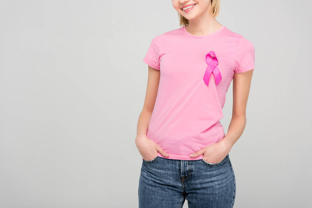 abgeschnittene Ansicht einer lächelnden jungen Frau in rosa T-Shirt mit Brustkrebs-Bewusstseinsband, isoliert auf grau - Foto, Bild