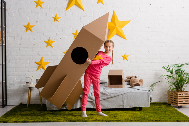 Niño dichoso en ropa rosa sosteniendo cohete de cartón grande en el dormitorio
 - Foto, imagen