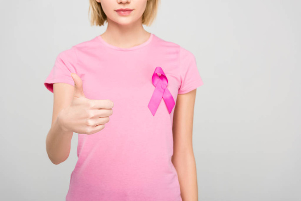 cropped view of young woman in pink t-shirt with breast cancer awareness ribbon showing thumbs up, isolated on grey  - Photo, Image
