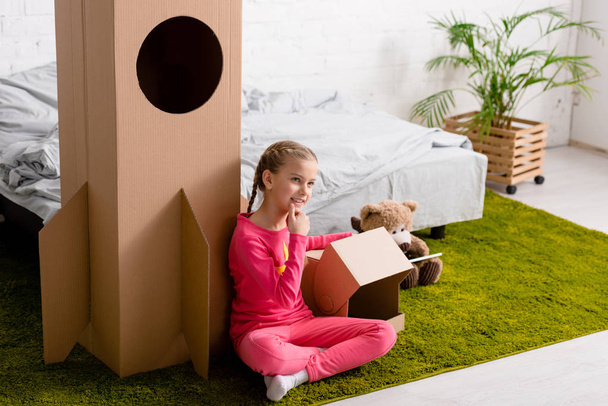 Curious kid in pink clothes sitting on carpet near cardboard rocket - Foto, afbeelding