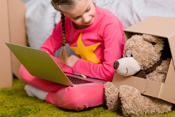 Cheerful kid with laptop sitting on carpet and looking at teddy bear - Valokuva, kuva