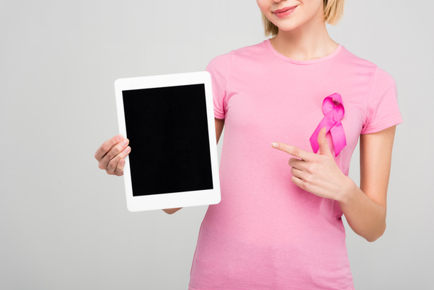 обрезанный вид девушки в розовой футболке с лентой осведомленности о раке молочной железы, указывая на планшет с пустым экраном, изолированный на сером
 - Фото, изображение