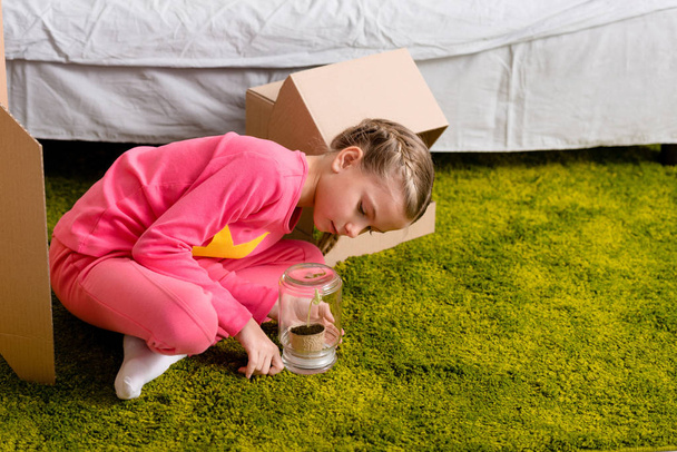 Kid sitting on green carpet and looking at plant in glass jar - Photo, Image