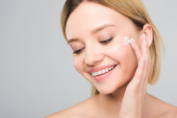 beautiful smiling young woman applying cosmetic cream on face isolated on grey - Fotografie, Obrázek