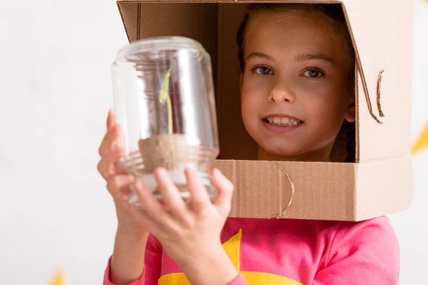 Ενθουσιασμένος παιδί στο χαρτόνι κράνος, κρατώντας το φυτό σε βάζο - Φωτογραφία, εικόνα