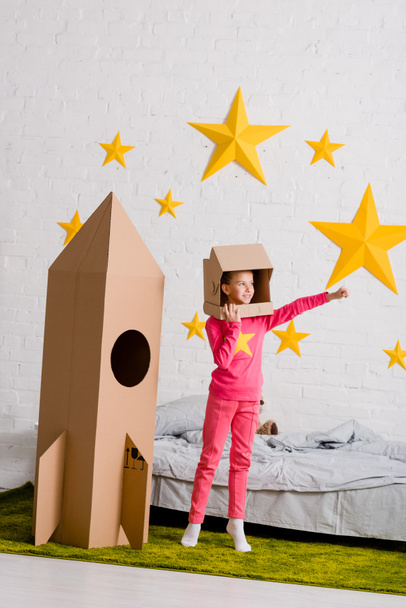 Full length view of kid in cardboard helmet standing near rocket in bedroom - Foto, immagini