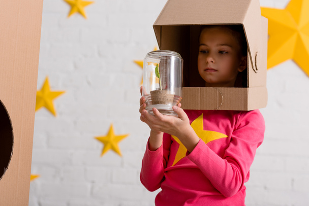 Curious child in cardboard helmet holding jar with plant - Fotoğraf, Görsel