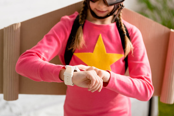 abgeschnittene Ansicht eines Kindes mit Pappflügeln, das auf die Uhr schaut - Foto, Bild