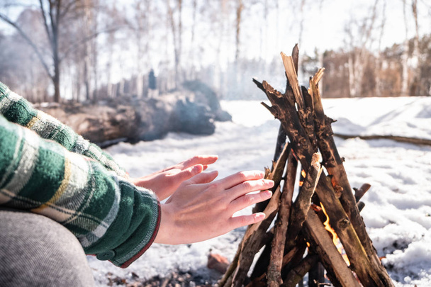 Basking near a campfire in a snowy birch forest. Female person getting warm near a fire on a sunny winter day in the woods - Фото, зображення