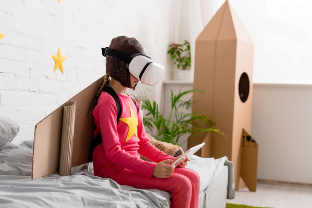 Kid in vr headset sitting on bed and using digital tablet - Фото, изображение