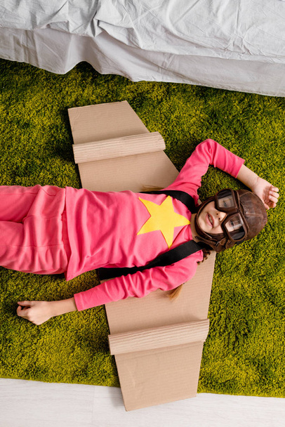 Top view of adorable kid with cardboard wings lying on green carpet - Foto, immagini