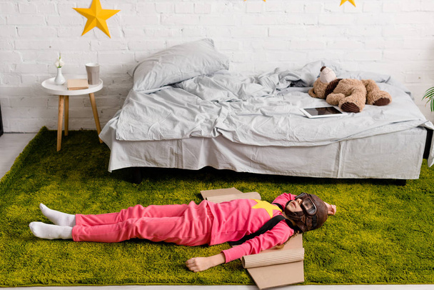 Niño cansado con alas de cartón acostado en la alfombra en el dormitorio
 - Foto, Imagen
