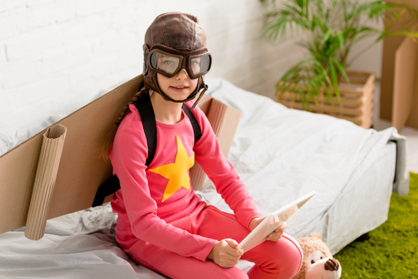 Niño curioso en casco de vuelo sentado en la cama y sosteniendo la tableta digital
 - Foto, Imagen