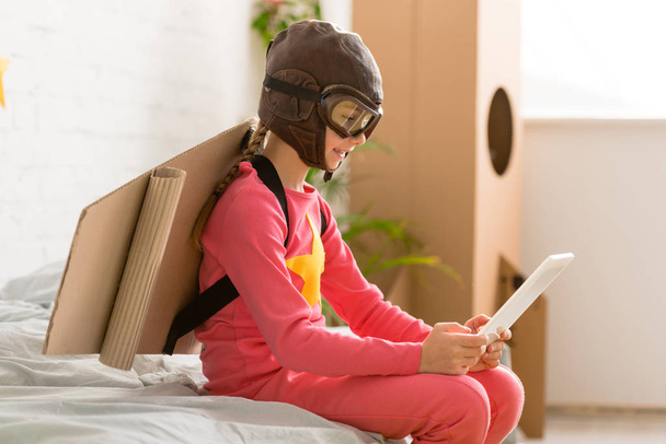 entzückendes Kind mit Pappflügeln auf dem Bett sitzend und mit digitalem Tablet - Foto, Bild
