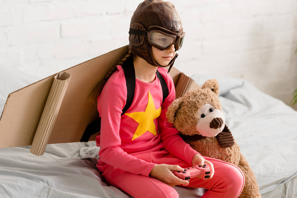 Kind met teddy beer zittend op bed en houden van de joystick - Foto, afbeelding