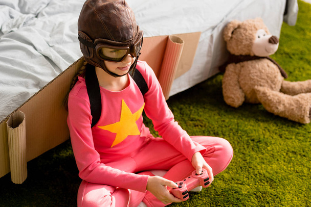 Niño con alas de cartón sentado en la alfombra y jugando videojuego con joystick
 - Foto, Imagen
