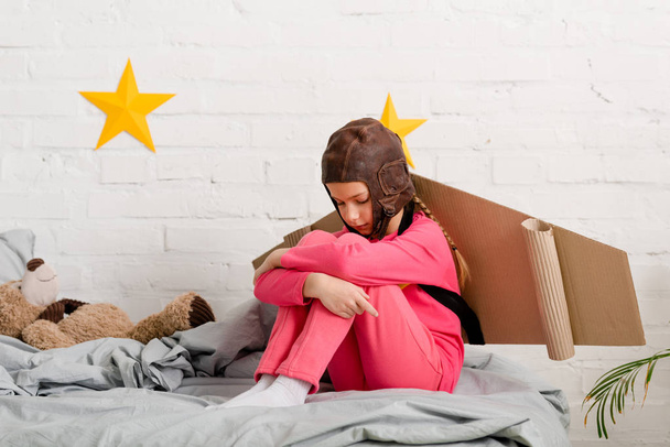 Üzgün çocuk yatağa oturmuş ve diz kucaklayan karton kanatlı - Fotoğraf, Görsel