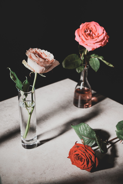 selective focus of pink and red rose flowers in glass bottles on stone table isolated on black - Photo, Image