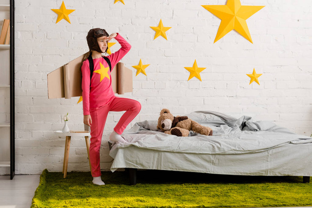 Excited kid with cardboard wings standing in bedroom and looking away - Foto, imagen