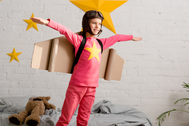 Niño inspirado con alas de cartón haciendo gestos en el dormitorio
 - Foto, Imagen
