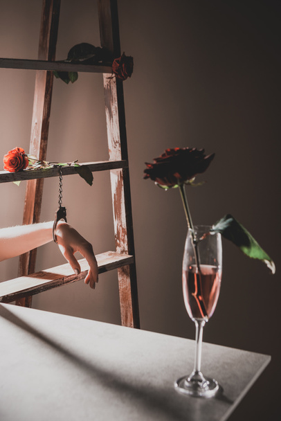 vermelho rosa flores em taça de champanhe na mesa de pedra com escada de madeira e mulher em algemas de metal no fundo
 - Foto, Imagem