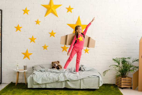 Blissful kid with cardboard wings standing on bed with hand up - Foto, Bild