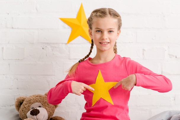 Cheerful kid with braids pointing with fingers at yellow star - Foto, imagen