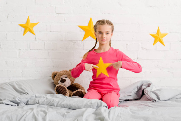 Παιδί με πλεξούδες κάθεται στο κρεβάτι και δείχνοντας με τα δάχτυλα στο κίτρινο αστέρι - Φωτογραφία, εικόνα