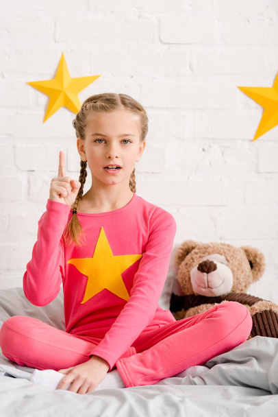 Pretty kid with teddy bear sitting on bed and holding finger up - Фото, зображення