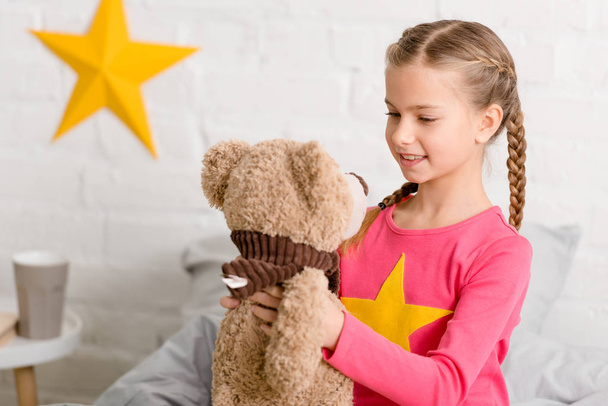 Blissful kid with braids looking at teddy bear with smile - Zdjęcie, obraz