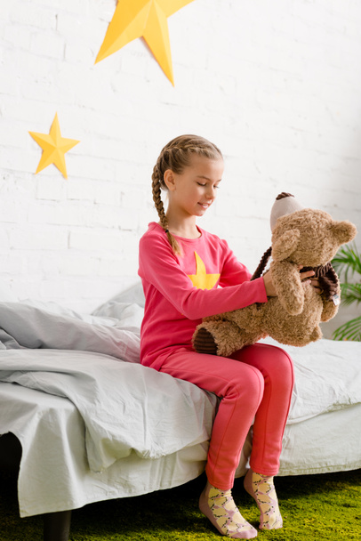 Cute kid with braids sitting on bed and looking at teddy bear - Фото, изображение