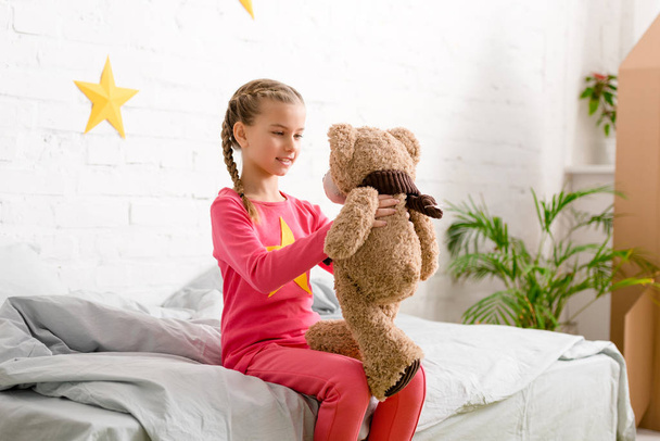 Charming kid with braids sitting on bed and looking at teddy bear - Photo, Image