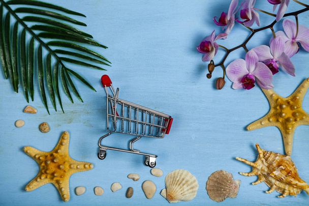 Planning a vacation at sea. Shopping cart, seashells, starfish and leaf of palm tree on a blue wooden background. - Photo, Image