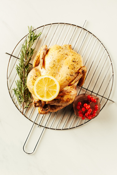 top view of fried chicken, rosemary and berries on metal grille with lemon on white table - Photo, image