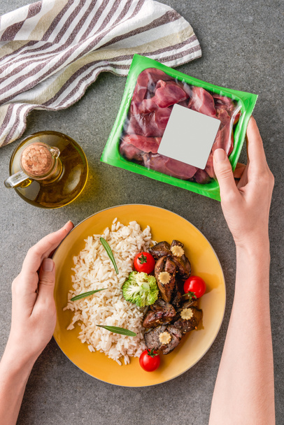 cropped view of woman holding plate with rice, meat and broccoli on plate with raw meat  - Foto, afbeelding