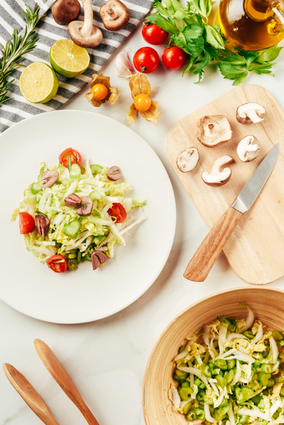 salade, chou et céleri en assiette avec bouteille d'huile, tomates cerises, chaux et champignons sur planche à découper
  - Photo, image