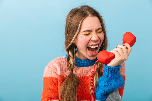 Excited young girl wearing winter clothes standing isolated over blue background, talking on a landline phone - Fotó, kép