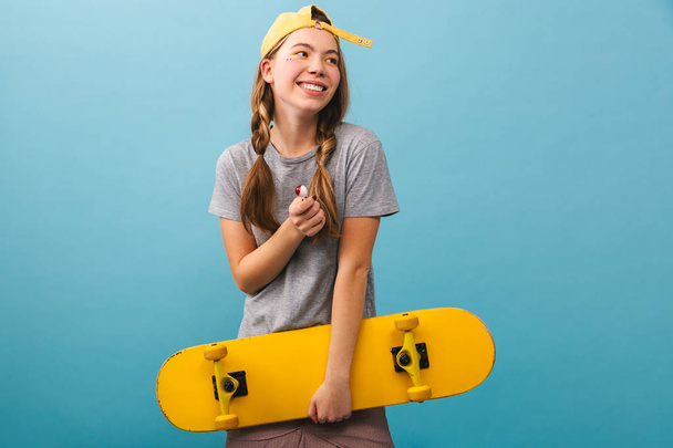 Cheerful cute girl standing isolated over blue background, holding skateboard - Valokuva, kuva