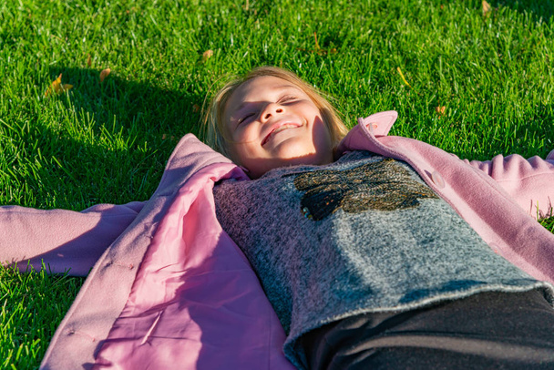Het meisje in de roze jas ligt op het groene gras in het park in het najaar - Foto, afbeelding
