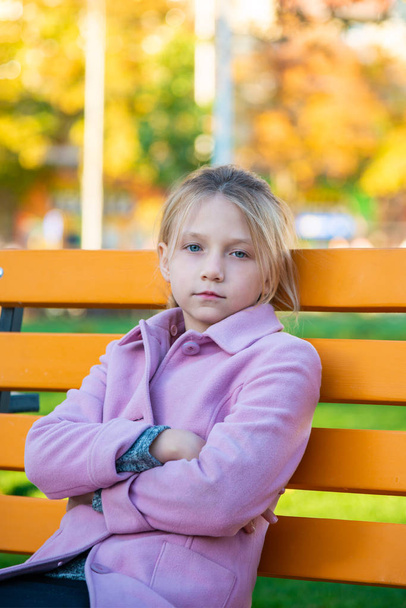 Vaaleanpunainen takki päällä oleva tyttö taittoi kätensä eteen ja katsoi kameraan istuen keltaisella penkillä puistossa.
 - Valokuva, kuva