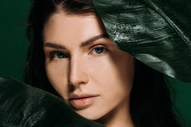 attractive woman with freckles on face posing with palm leaves isolated on green - Foto, Imagem