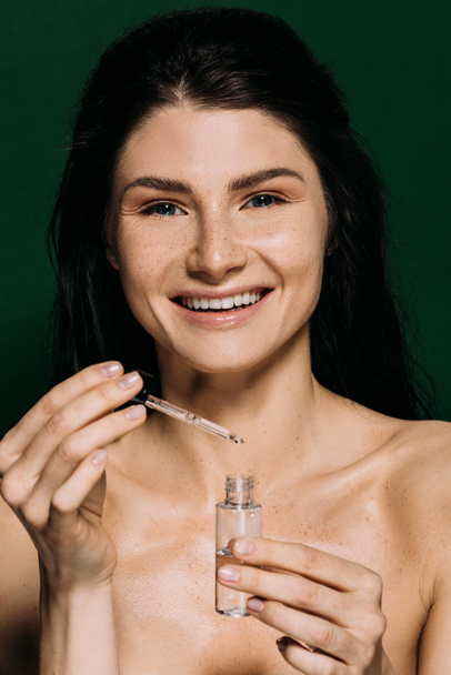 cheerful naked girl holding bottle with moisturizing serum isolated on green - Foto, afbeelding