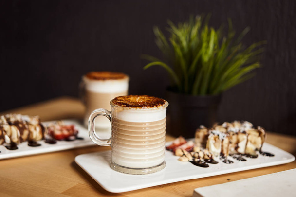 Two cups of Hot latte with baked caramel crust and sweet roll with banana and strawberry on the wooden table in coffe-shop. Coffee concept. - Фото, изображение