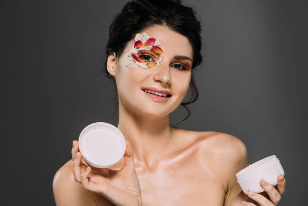 beautiful woman with petals on face holding plastic container with moisturizing cream isolated on grey - Photo, Image
