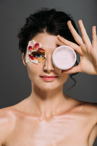 schöne Frau mit Blütenblättern im Gesicht hält Plastikbehälter mit kosmetischer Creme isoliert auf grau - Foto, Bild