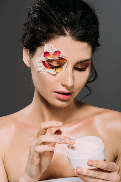 attractive young woman with petals on face holding plastic container with cream isolated on grey - Foto, Imagen